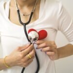Unrecognizable female wearing white shirt while standing on white background with diaphragm of stethoscope on red handmade heart in room