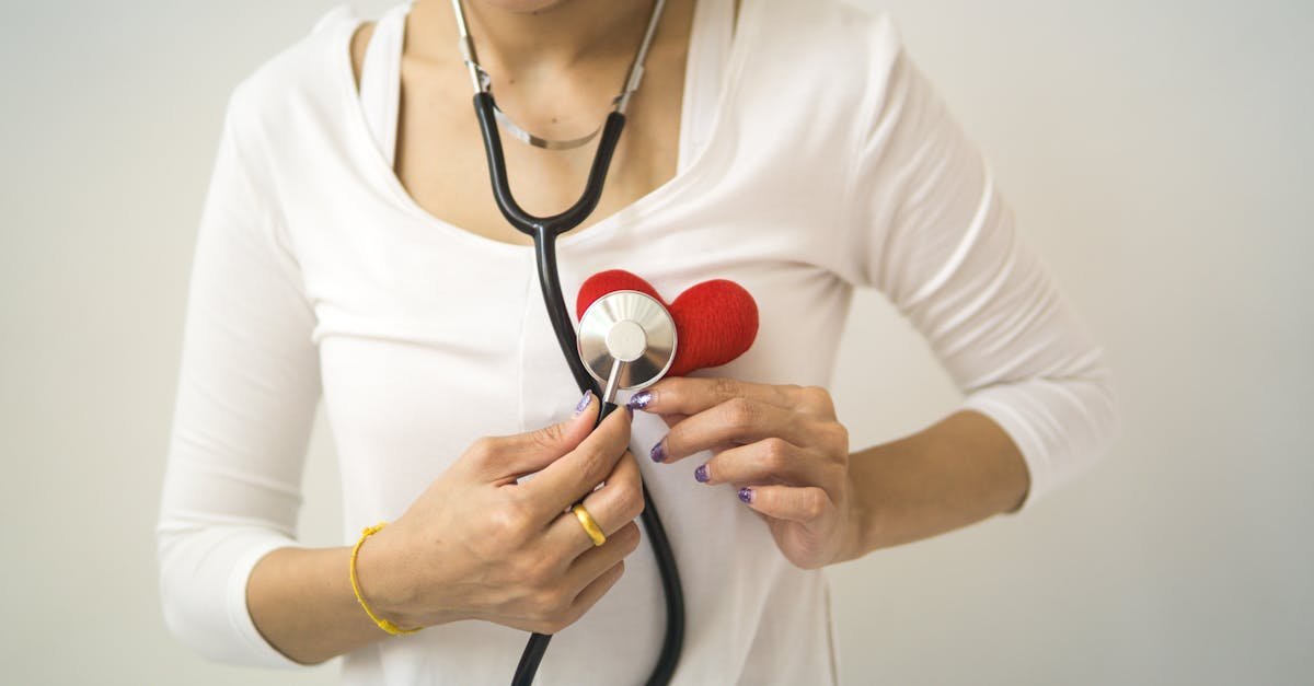 Unrecognizable female wearing white shirt while standing on white background with diaphragm of stethoscope on red handmade heart in room