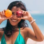 Woman at the beach smiling while playfully holding an apple and orange, embodying summer fun.