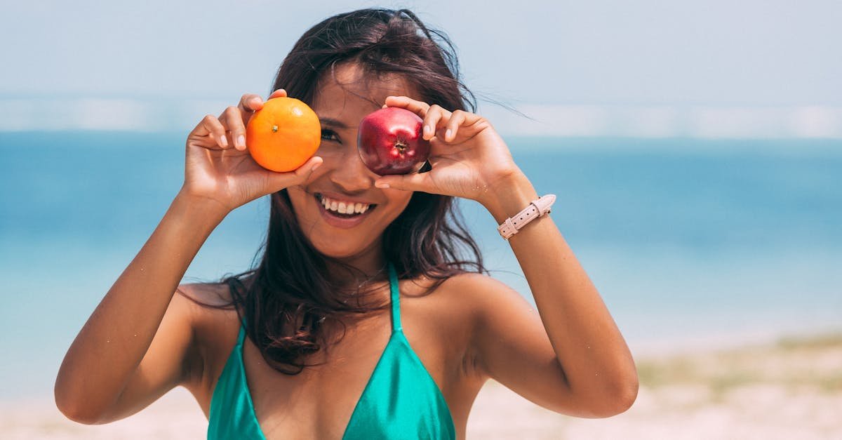 Woman at the beach smiling while playfully holding an apple and orange, embodying summer fun.