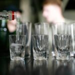 Close-up of a bottle pouring clear alcohol into shot glasses, with people in the background.