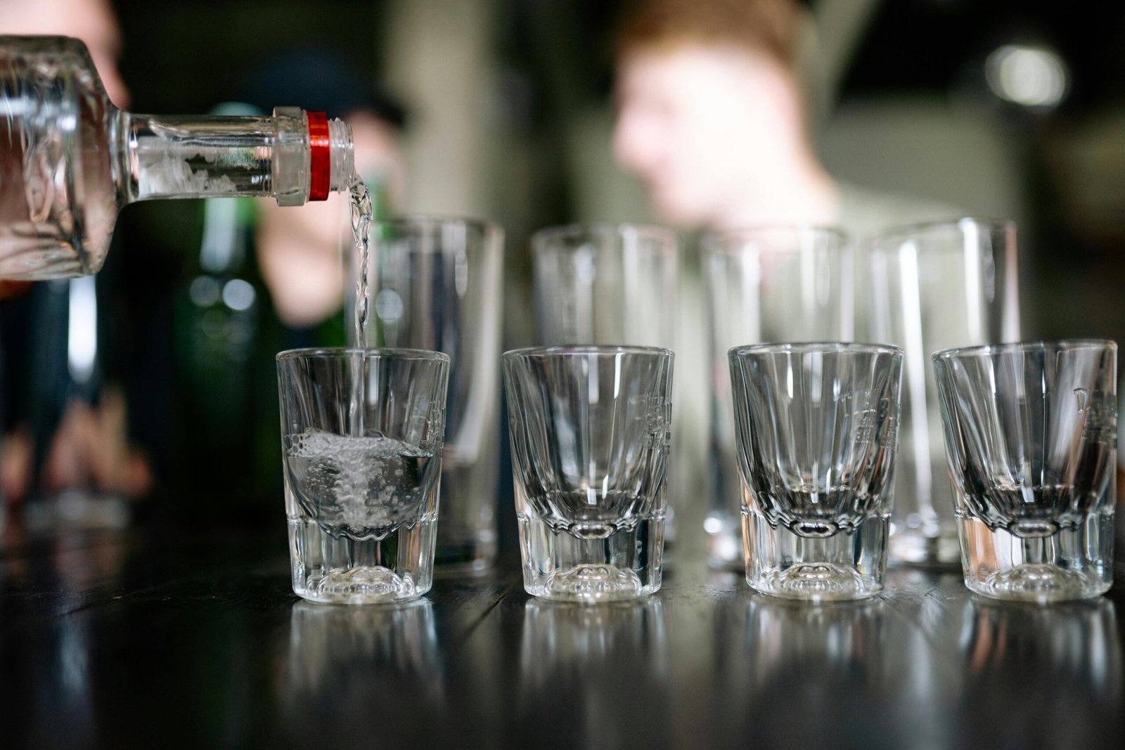 Close-up of a bottle pouring clear alcohol into shot glasses, with people in the background.
