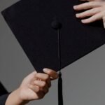 Two people exchanging a graduation cap on a grey background, symbolizing achievement.