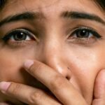 Close-up portrait of a woman with hand covering mouth, showing a scared expression, in a studio setting.