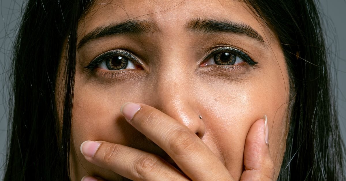 Close-up portrait of a woman with hand covering mouth, showing a scared expression, in a studio setting.