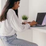 Young woman in a white shirt working on a laptop at a minimalist home office desk.