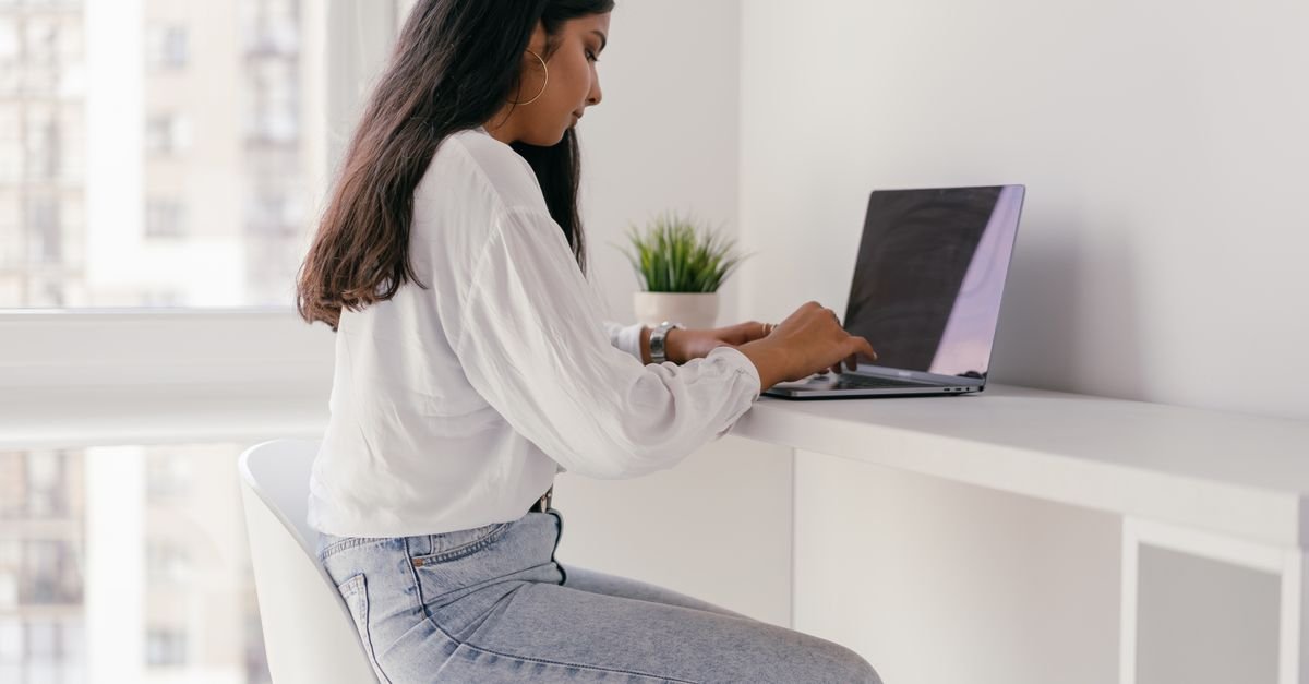 Young woman in a white shirt working on a laptop at a minimalist home office desk.
